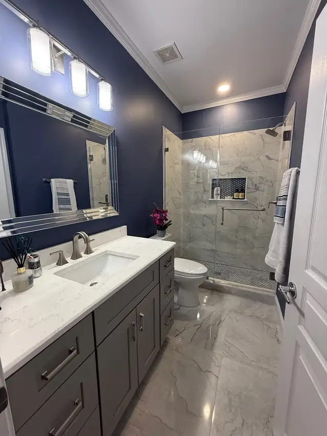 Remodeled bathroom with navy walls, a gray vanity with a white top and a glass walk-in shower with marble-look tile