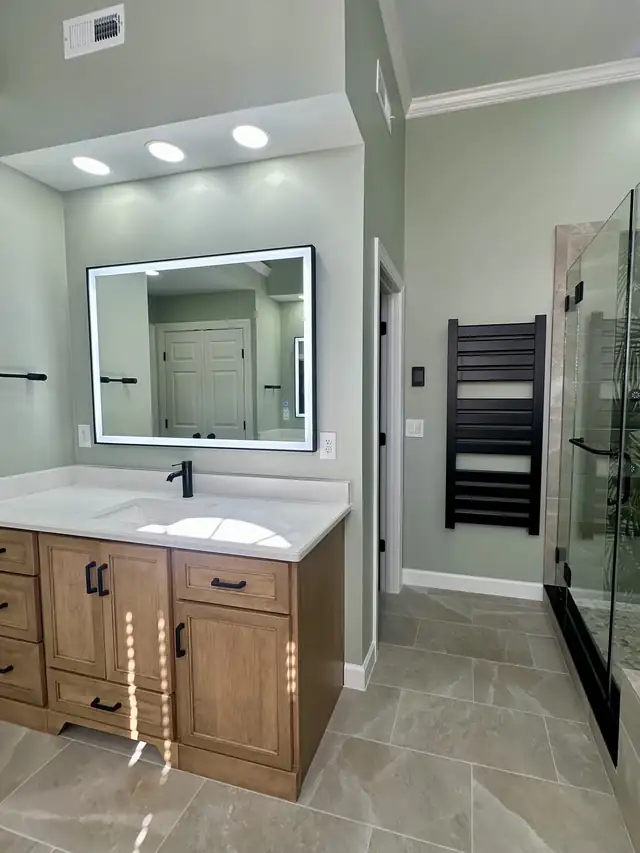 Wood bathroom vanity with white top, backlit mirror, black towel warmer and frameless shower in a remodeled Triangle bathroom