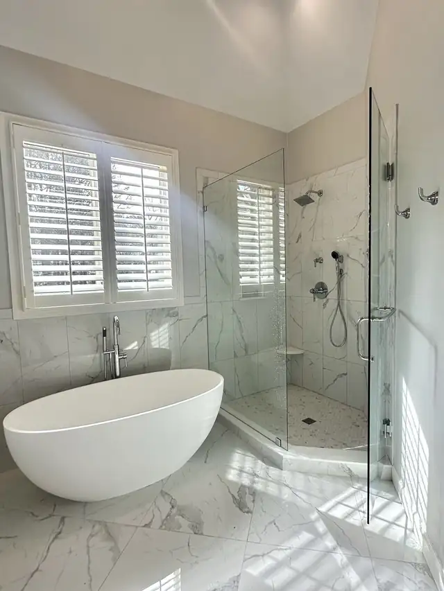 Freestanding white soaking tub next to a glass-enclosed shower with marble-pattern walls and plantation shutters in a remodeled bathroom